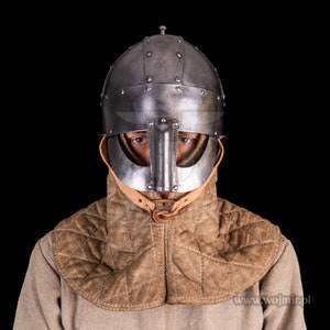 Puede incluir: Una persona con un casco medieval con protecci&oacute;n para la nariz y una protecci&oacute;n para el cuello acolchada de color marr&oacute;n. El casco est&aacute; hecho de metal y tiene un acabado oscuro. La protecci&oacute;n para el cuello est&aacute; hecha de tela y tiene un patr&oacute;n acolchado. La persona est&aacute; mirando directamente a la c&aacute;mara.