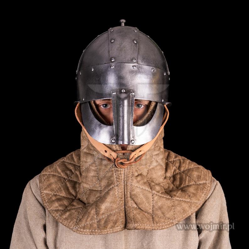 Puede incluir: Una persona con un casco medieval con protecci&oacute;n para la nariz y una protecci&oacute;n para el cuello acolchada de color marr&oacute;n. El casco est&aacute; hecho de metal y tiene un acabado oscuro. La protecci&oacute;n para el cuello est&aacute; hecha de tela y tiene un patr&oacute;n acolchado. La persona est&aacute; mirando directamente a la c&aacute;mara.