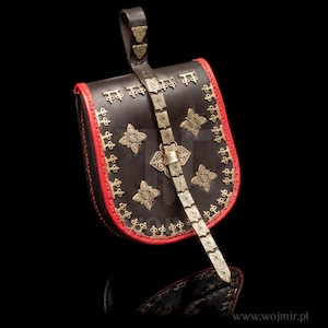 Pode incluir: Uma bolsa de couro marrom com acabamento vermelho e detalhes em metal dourado. A bolsa tem uma alça com fivela e é decorada com padrões intrincados.