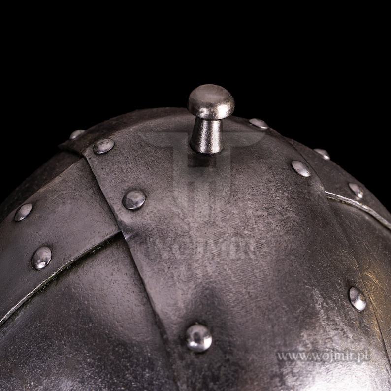 Puede incluir: Primer plano de la parte superior de un casco plateado con un pomo redondo en la parte superior. El casco est&aacute; hecho de placas de metal que est&aacute;n remachadas juntas. El casco tiene un aspecto desgastado y envejecido.