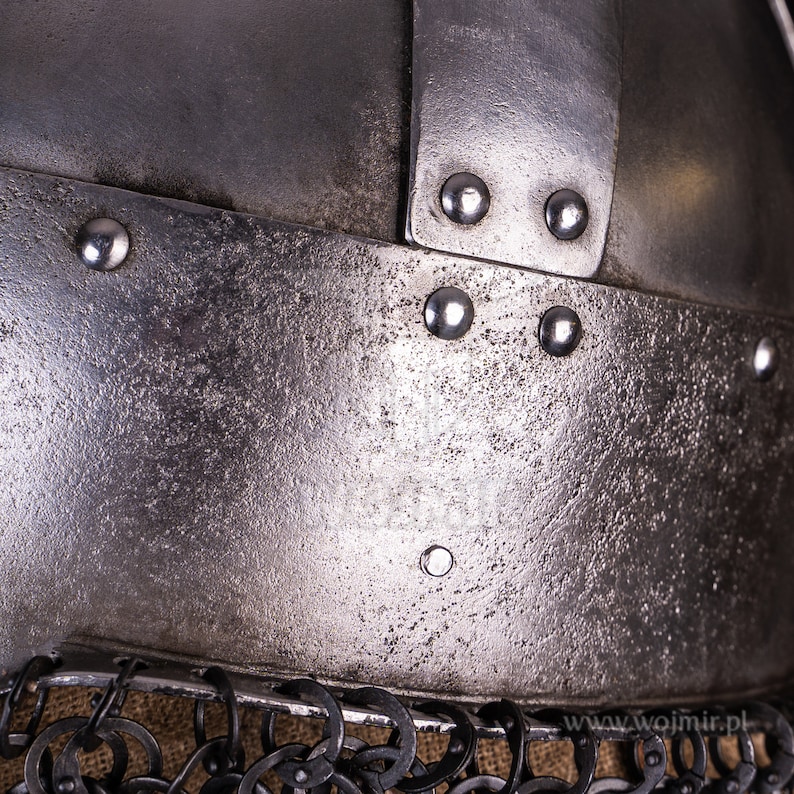 Puede incluir: Primer plano de un casco plateado con remaches y una cota de malla. El casco tiene una superficie texturizada y un peque&ntilde;o remache redondo en el centro.