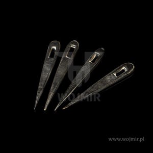 Puede incluir: Cuatro agujas de tejer de metal plateado con puntas puntiagudas y agujeros para los ojos. Las agujas están dispuestas en forma de abanico sobre un fondo negro.