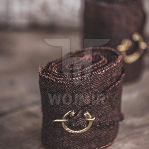 May include: Two rolled brown fabric pieces with a woven pattern, each secured with a gold-colored clasp. The fabric has a reddish-brown hue. The items are on a wooden surface.