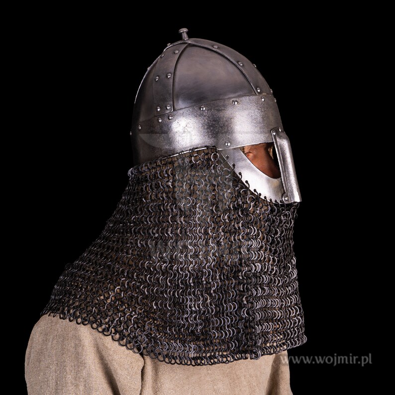 Puede incluir: Un casco de plata con una cota de malla, usado por una persona. El casco tiene una parte superior puntiaguda y una m&aacute;scara facial con una peque&ntilde;a abertura para los ojos. La cota de malla cubre el cuello y los hombros.