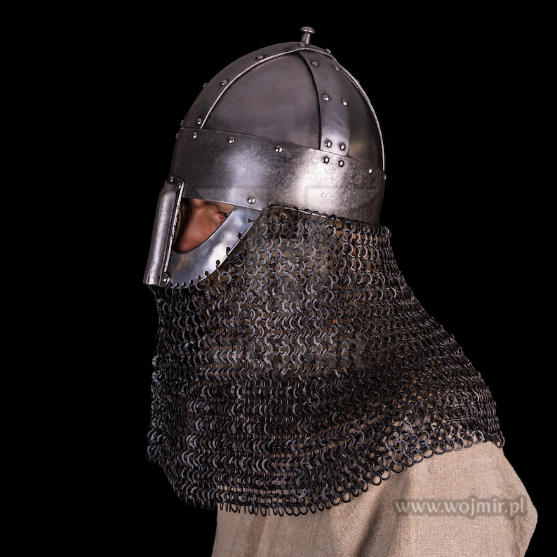 Puede incluir: Un casco de plata con una cota de malla, usado por una persona. El casco tiene una protecci&oacute;n de nariz puntiaguda y una parte superior redondeada. La cota de malla cubre el cuello y los hombros.