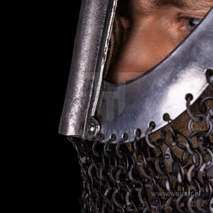 Puede incluir: Primer plano de un casco plateado con una m&aacute;scara de malla de cadena. El casco tiene una visera que est&aacute; parcialmente abierta, revelando el ojo de la persona que lo lleva. El casco est&aacute; hecho de metal y tiene una superficie texturizada. La malla de cadena est&aacute; hecha de peque&ntilde;os anillos de metal que est&aacute;n unidos entre s&iacute;. El casco es una pieza de armadura medieval.