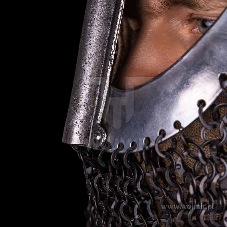 Puede incluir: Primer plano de un casco plateado con una m&aacute;scara de malla de cadena. El casco tiene una visera que est&aacute; parcialmente abierta, revelando el ojo de la persona que lo lleva. El casco est&aacute; hecho de metal y tiene una superficie texturizada. La malla de cadena est&aacute; hecha de peque&ntilde;os anillos de metal que est&aacute;n unidos entre s&iacute;. El casco es una pieza de armadura medieval.