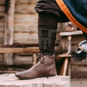 May include: Brown leather boots with laces and a brown fabric cuff. The boots are worn by a person standing on a wooden surface.