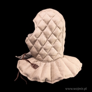 Puede incluir: Cofia acolchada beige, un tipo de tocado medieval, con un patrón de diamantes. La cofia tiene un borde inferior con volantes y una etiqueta de cuero con una cinta marrón. El fondo es negro.