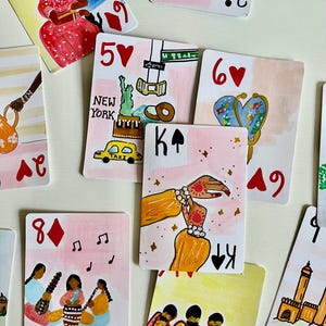 Könnte beinhalten: Eine Sammlung handbemalter Spielkarten mit verschiedenen Illustrationen, darunter eine Person in einem roten Sari, eine New Yorker Szene mit Taxi und Freiheitsstatue, Hände mit Hennamustern, musizierende Personen und architektonische Wahrzeichen.