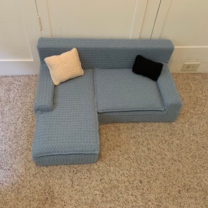 May include: A light blue sectional sofa with a white pillow and a black pillow. The sofa is on a beige carpet.