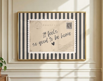 May include: A framed wall art print featuring a vintage-style postcard design with the text "It feels so good to be home" and a small heart icon. The postcard is centered on a background of vertical black and white stripes within a gold frame.