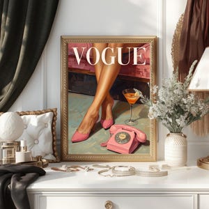 May include: A framed wall art print featuring the word VOGUE in white capital letters. The illustration depicts a pair of legs in pink high-heeled shoes, a martini glass, and a vintage pink rotary telephone on a light green floor surface.