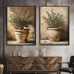 May include: Two framed paintings of olive trees in terracotta pots against a beige background. The paintings are in black frames and are hung on a gray wall.