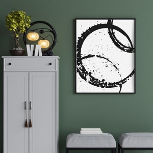 May include: A white cabinet with two doors and a drawer, a black metal lamp with three light bulbs, a small potted tree, and a black and white abstract painting with a black frame hanging on a green wall.