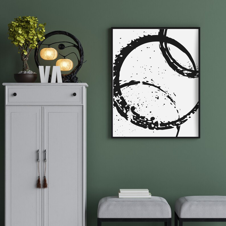 May include: A white cabinet with two doors and a drawer, a black metal lamp with three light bulbs, a small potted tree, and a black and white abstract painting with a black frame hanging on a green wall.