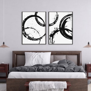 May include: Two black and white abstract art prints with black circles and splatters on a white background. The prints are framed in black and hung on a white wall above a gray bed with white bedding.