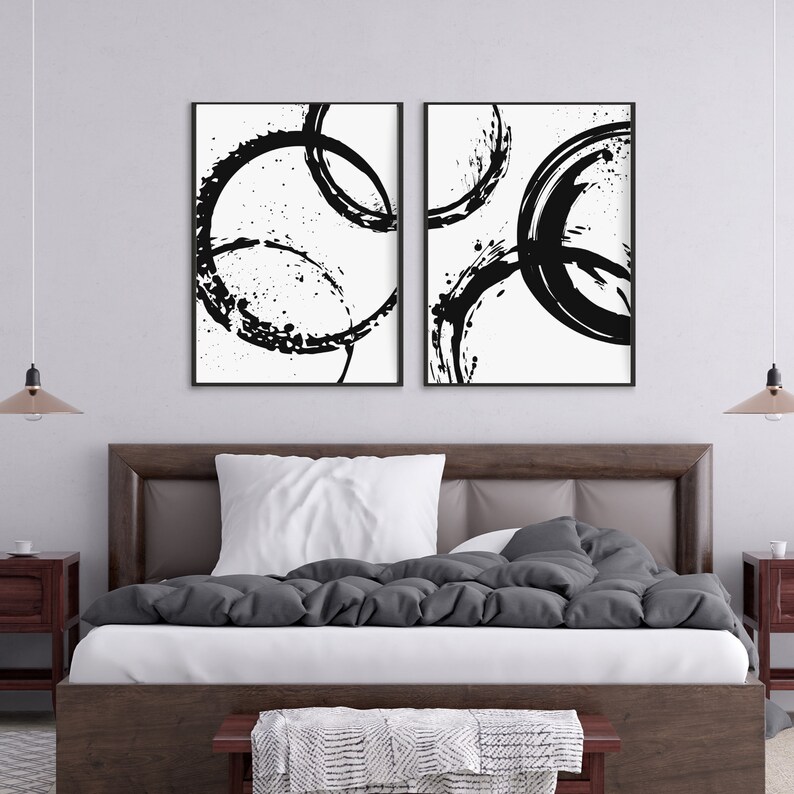 May include: Two black and white abstract art prints with black circles and splatters on a white background. The prints are framed in black and hung on a white wall above a gray bed with white bedding.