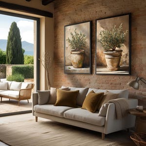 May include: A light beige sofa with two brown throw pillows sits in a living room with a large window overlooking a green landscape. Two framed paintings of potted olive trees hang on a brick wall behind the sofa.