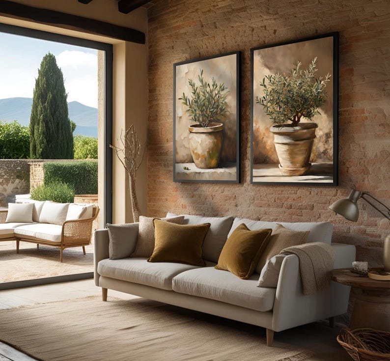 May include: A light beige sofa with two brown throw pillows sits in a living room with a large window overlooking a green landscape. Two framed paintings of potted olive trees hang on a brick wall behind the sofa.