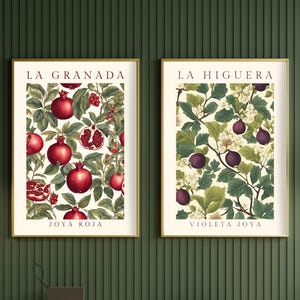 May include: Two framed botanical prints with gold frames. The print on the left features a pomegranate branch with red pomegranates and green leaves. The print on the right features a fig branch with purple figs and green leaves. The print on the left has the text "LA GRANADA" at the top and "JOYA ROJA" at the bottom. The print on the right has the text "LA HIGUERA" at the top and "VIOLETA JOYA" at the bottom.