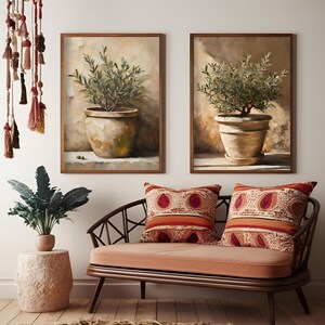 May include: Two framed paintings of potted olive trees. The paintings are in a modern style with a neutral color palette. The paintings are hanging on a white wall above a brown couch with two red and orange patterned pillows.