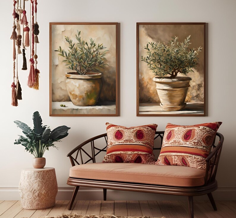 May include: Two framed paintings of potted olive trees. The paintings are in a modern style with a neutral color palette. The paintings are hanging on a white wall above a brown couch with two red and orange patterned pillows.