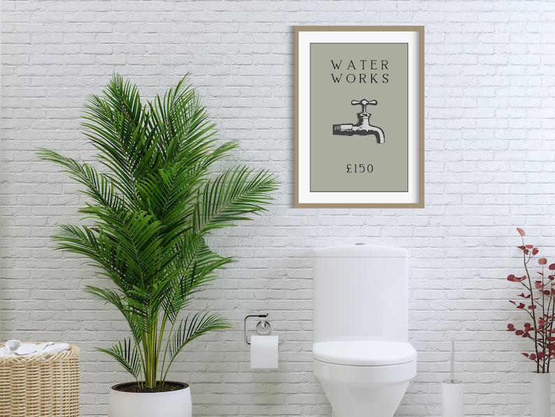 May include: A framed wall art print featuring a sage green background with the black text WATER WORKS above a black and white illustration of a water faucet and the text &pound;150 below it, all enclosed in a simple light brown wooden frame.