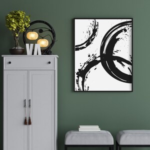 May include: A white cabinet with two doors and a drawer, a black metal light fixture with three round light bulbs, a small potted plant, and a black framed abstract painting with black brushstrokes on a white background.