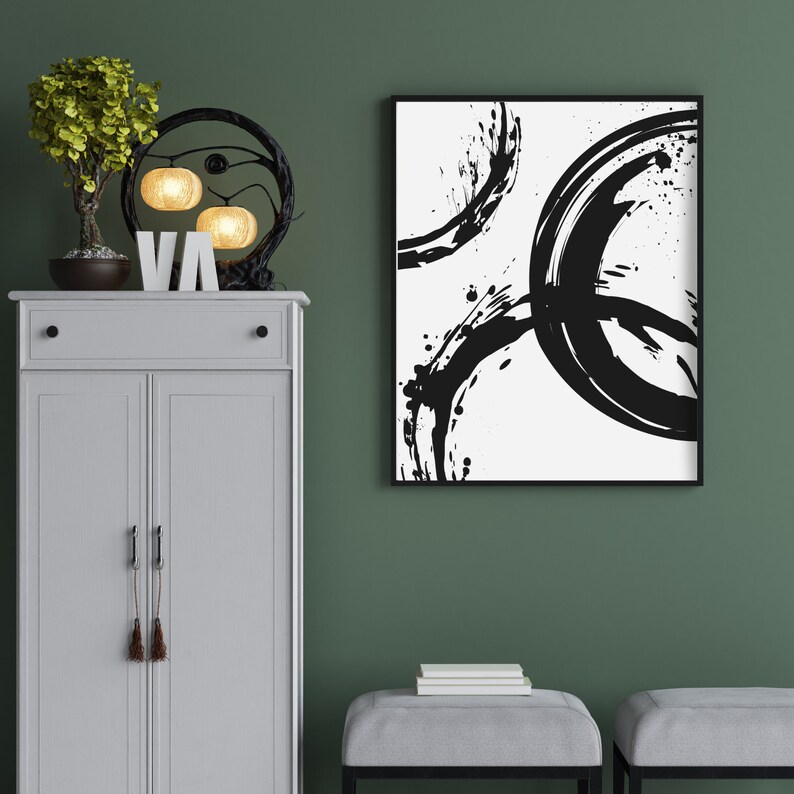 May include: A white cabinet with two doors and a drawer, a black metal light fixture with three round light bulbs, a small potted plant, and a black framed abstract painting with black brushstrokes on a white background.