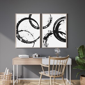 May include: Two black and white abstract art prints with a circular design. The prints are framed in a light wood frame and are hanging on a gray wall.