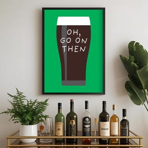 Puede incluir: Un cuadro enmarcado con una pinta de cerveza oscura y el texto "OH, GO ON THEN" sobre un fondo verde. El cuadro est&aacute; en un marco negro. Un carrito bar con varias botellas de vino y licores est&aacute; en primer plano.