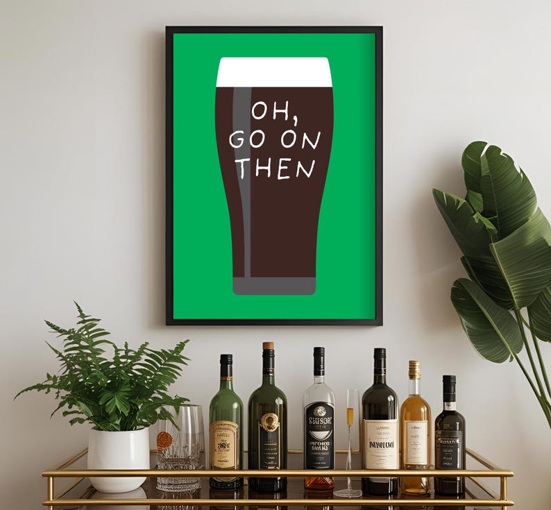 Puede incluir: Un cuadro enmarcado con una pinta de cerveza oscura y el texto "OH, GO ON THEN" sobre un fondo verde. El cuadro est&aacute; en un marco negro. Un carrito bar con varias botellas de vino y licores est&aacute; en primer plano.