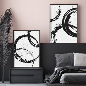 May include: Two black and white abstract art prints with circular brushstrokes on a white background. The prints are framed in black and hung on a wall above a black nightstand and a bed with gray bedding.