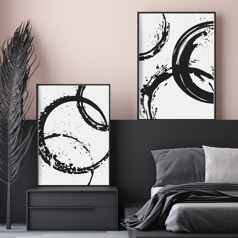 May include: Two black and white abstract art prints with circular brushstrokes on a white background. The prints are framed in black and hung on a wall above a black nightstand and a bed with gray bedding.