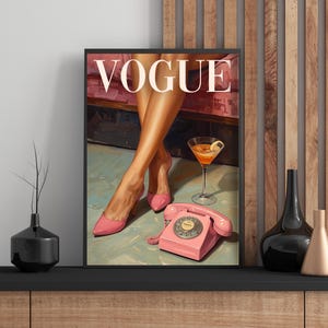 May include: A framed wall art print featuring the word VOGUE in white capital letters. The illustration depicts a pair of legs in pink high-heeled shoes, a pink rotary telephone, and a cocktail glass with a lemon slice on a light surface.