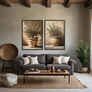 May include: Two framed paintings of potted olive trees with brown pots and green leaves against a beige wall. The paintings are hanging above a gray couch with pillows and a wooden coffee table.