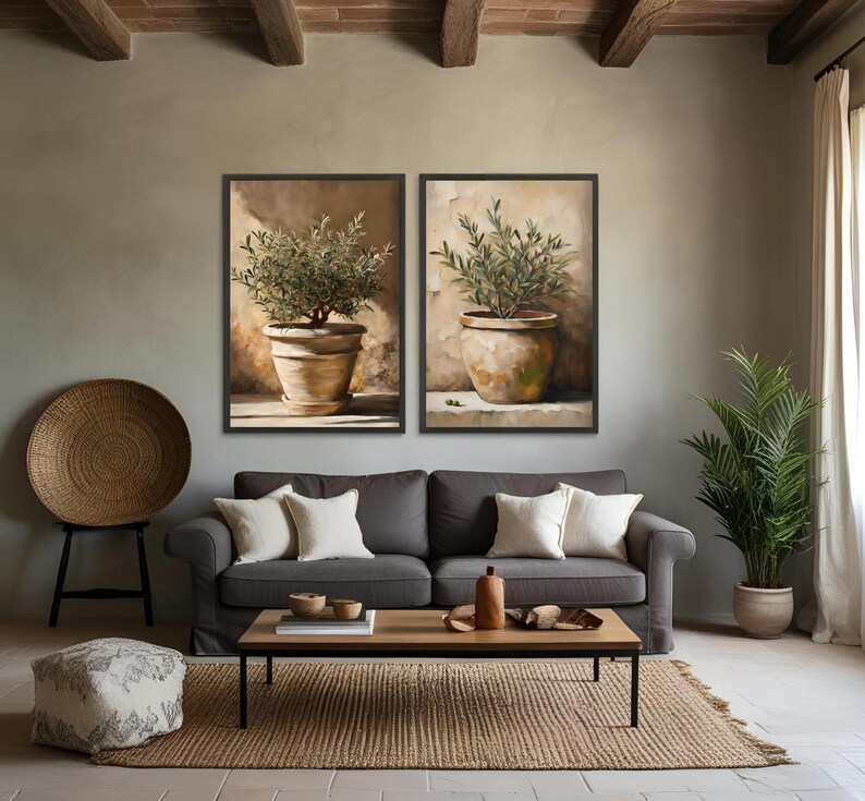 May include: Two framed paintings of potted olive trees with brown pots and green leaves against a beige wall. The paintings are hanging above a gray couch with pillows and a wooden coffee table.
