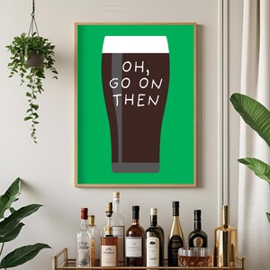 Puede incluir: Un cuadro enmarcado con una pinta de cerveza oscura y el texto "OH, GO ON THEN" en blanco. El fondo es verde y el marco es de madera clara. El cuadro se muestra sobre un carrito de bar con varias botellas.