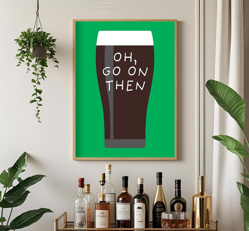 Puede incluir: Un cuadro enmarcado con una pinta de cerveza oscura y el texto "OH, GO ON THEN" en blanco. El fondo es verde y el marco es de madera clara. El cuadro se muestra sobre un carrito de bar con varias botellas.