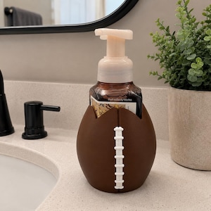 May include: A brown soap dispenser holder shaped like an American football with white laces, containing a Bath & Body Works foaming soap bottle with a light-colored pump dispenser. The holder is designed to fit around the base of the soap bottle.