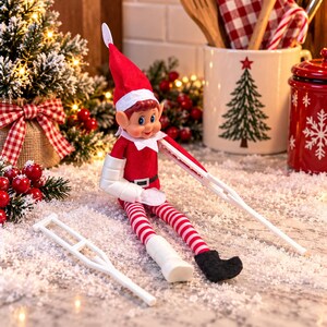 May include: A plush elf doll wearing a red hat and suit with a white collar and belt. The doll has a white cast on one arm and one leg, and is using white crutches. A second white crutch lies on the surface nearby. The doll has red and white striped legs.