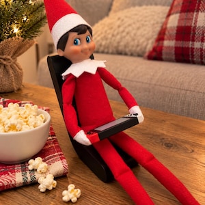 May include: A plush elf doll with a red suit, white collar, and red pointed hat sits in a black chair. The doll holds a small handheld gaming console with red and blue controllers. A white bowl of popcorn sits nearby on a wooden surface.