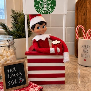 May include: A miniature Elfbucks coffee stand featuring a red and white striped base, a white roof with a green circular logo, and a doll wearing a red and white outfit holding a red mug with marshmallows. A small chalkboard sign reads Hot Cocoa 25¢.