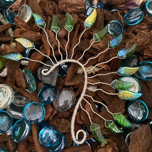 Puede incluir: Un brazalete de oreja de alambre plateado con un diseño de hojas iridiscentes verdes y azules. El brazalete se muestra sobre una cama de virutas de madera marrón y piedras de vidrio azules.