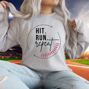 May include: A light gray sweatshirt with the text "HIT. RUN. repeat" in black and a pink baseball graphic. The sweatshirt is a crew neck style and is made of a soft material.