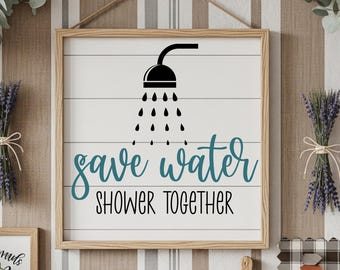 Puede incluir: Cartel enmarcado en madera con fondo blanco y lamas horizontales de madera. El cartel presenta un gráfico de alcachofa de ducha negra con gotas de agua y el texto turquesa "save water" sobre "shower together" en negro.