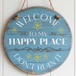 Może przedstawiać: Okrągły, niebieski drewniany znak z napisem "WELCOME TO MY HAPPY PLACE DON'T RUIN IT" w białych literach. Znak ma żółte kwiatowe akcenty i sznurek do zawieszenia. Drewno ma postarzaną powierzchnię.