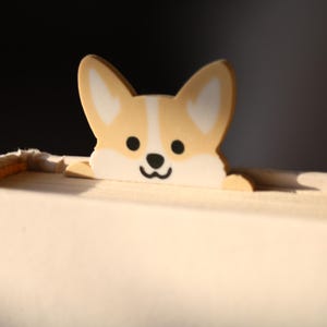Könnte beinhalten: Ein Lesezeichen in Form eines Corgi-Hundes, beige und weiß, liegt auf einem Buch. Das Lesezeichen hat ein lächelndes Gesicht mit schwarzen Augen und einer schwarzen Nase. Die Seiten des Buches sind cremefarben.