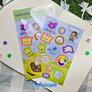 May include: Two sticker sheets with colorful cartoon characters and the text "welcome to kinzville!" are displayed on a white notebook. The stickers feature various designs, including crowns, animals, and a color wheel. Scattered around are star-shaped embellishments.